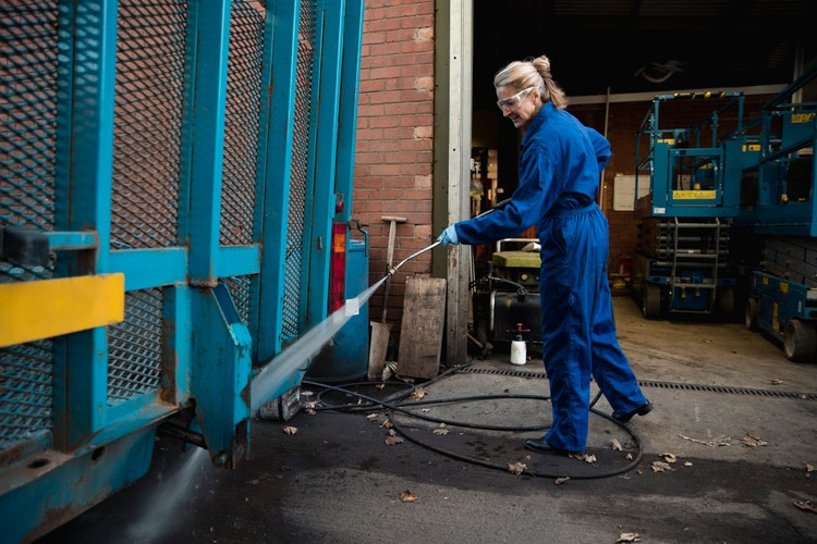A side-view shot of a female manual worker cleaning a vehicle exterior with a high-pressure​ water hose.